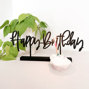 Happy Birthday Little Word Stand painted black and standing on a white table with a neon pathos plant in the background and a cupcake with a lit rainbow candle in the front