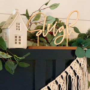 The phrase "Sleigh" in natural wood standing on a blue mantel with holiday greenery behind