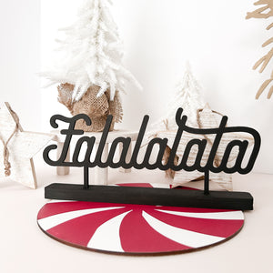 The word "Falalalala" cut out in a black cursive font standing in a black stand. It stands on a red and white peppermint plate and in the background are small white christmas trees and white washed wood stars