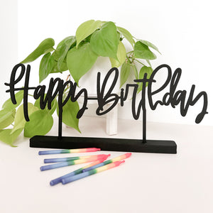 The phrase "Happy Birthday" in black is standing on a white background with rainbow birthday candles laying on the table. There is a neon pathos in the background.