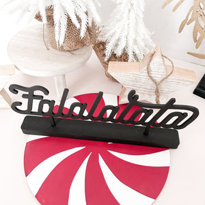 A top down view of the Falalalala word stand cut in black. In the background is white washed wood stars and plant stand that hold a small white christmas tree