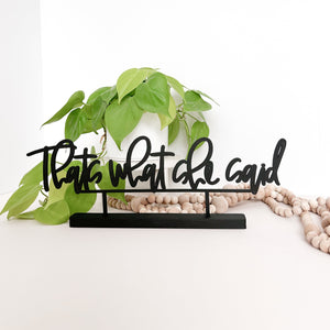 The phrase "thats what she said" in black standing on a little word stand holder on a white background. Neon pathos and wood bead garland in the back ground