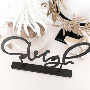 The word "sleigh" in black standing on a white background. Behind it is some wooden stars, a small white christmas tree and a gold wreath on a stand