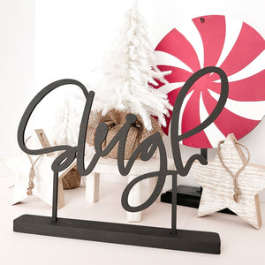 The word "sleigh" in black with a black stand. in the background is a peppermint candy on a stand, wooden stars and a small white christmas tree on a plant stand