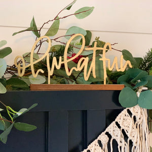 The phrase "oh what fun" in natural wood standing on a blue mantel with holiday greenery behind