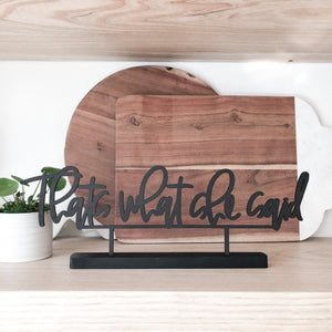 The phrase "thats what she said" cut out in black standing on a shelf. In the background there are wooden cutting boards and a baby pilea